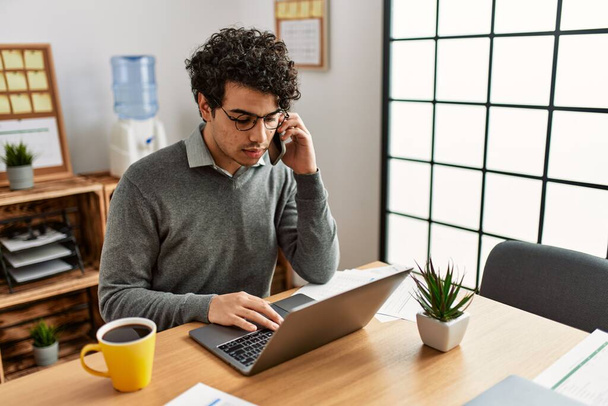 Молодой латиноамериканец разговаривает на смартфоне и пользуется ноутбуком в офисе. - Фото, изображение
