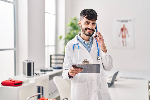 Young hispanic man wearing doctor uniform talking on smartphone reading medical report at clinic - Fotó, kép