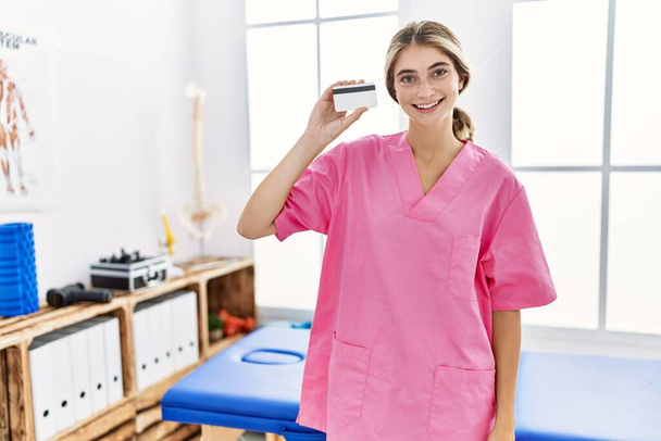 Mladá fyzioterapeutka žena pracující na klinice pro zotavení bolesti držící kreditní kartu vypadá pozitivně a šťastně vstoje a usmívá se s sebevědomým úsměvem ukazujícím zuby  - Fotografie, Obrázek