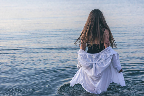 A woman in a swimsuit and a white shirt in the sea at sunset, view from the back. - Foto, Imagen