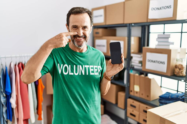 Middle age man with beard wearing volunteer t shirt holding smartphone pointing with hand finger to face and nose, smiling cheerful. beauty concept  - Foto, Imagem