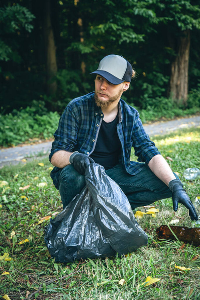 A young man with a garbage bag in the forest cleans up plastic bottles, the concept of love for nature and care for the ecology. - Photo, image