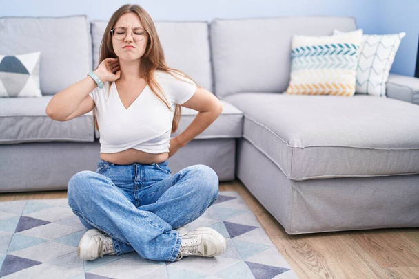 Young caucasian woman sitting on the floor at the living room suffering of neck ache injury, touching neck with hand, muscular pain  - Photo, Image