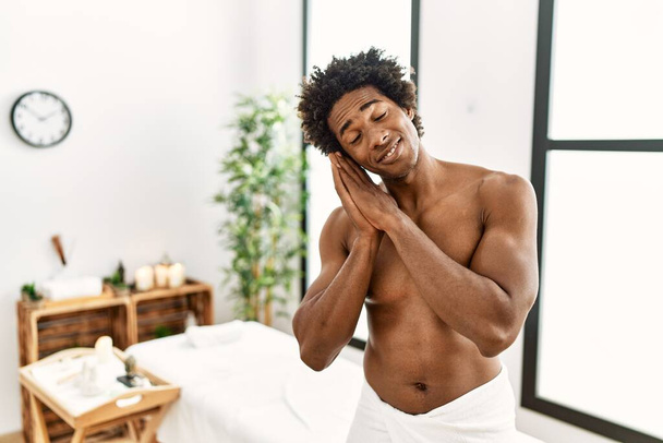 Genç Afrikalı Amerikalı üstsüz bir adam güzellik merkezinde havluyla dikiliyor. Yorgun uyuyor, hayal kuruyor ve elleri ile birlikte poz veriyor, gözleri kapalı gülümsüyor..  - Fotoğraf, Görsel