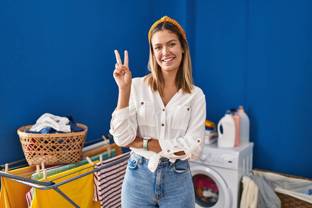 Young blonde woman at laundry room smiling with happy face winking at the camera doing victory sign with fingers. number two.  - Photo, image