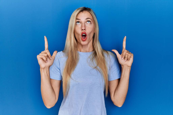 Όμορφη ξανθιά γυναίκα φορώντας casual t πουκάμισο πάνω από το μπλε φόντο έκπληκτος και κοιτάζοντας προς τα πάνω και δείχνοντας με τα δάχτυλα και σήκωσε τα χέρια.  - Φωτογραφία, εικόνα