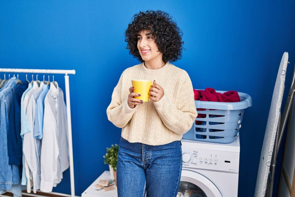Young middle east woman drinking coffee waiting for washing machine at laundry - 写真・画像