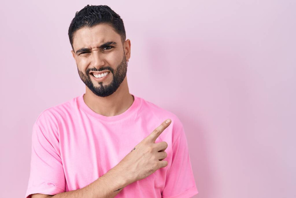 Ισπανόφωνος νεαρός άνδρας στέκεται πάνω από ροζ φόντο δείχνοντας στην άκρη ανήσυχος και νευρικός με δείκτη, ανησυχούν και έκπληκτος έκφραση  - Φωτογραφία, εικόνα
