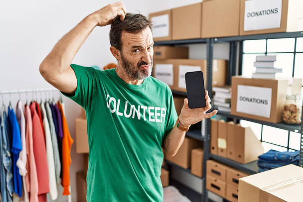 Middle age man with beard wearing volunteer t shirt holding smartphone confuse and wondering about question. uncertain with doubt, thinking with hand on head. pensive concept.  - Foto, Bild