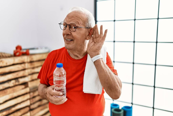 Senior man wearing sportswear and towel at the gym smiling with hand over ear listening an hearing to rumor or gossip. deafness concept.  - Foto, afbeelding