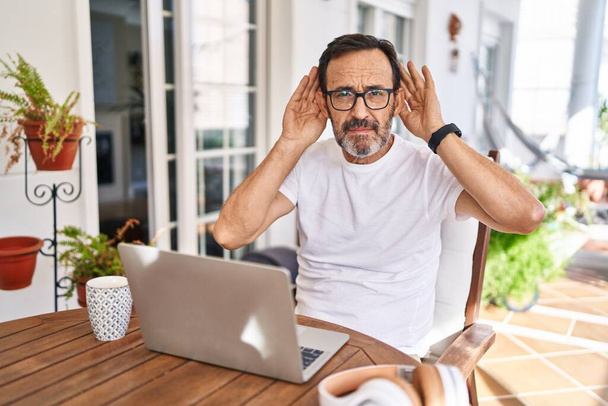 Middle age man using computer laptop at home trying to hear both hands on ear gesture, curious for gossip. hearing problem, deaf  - Foto, afbeelding