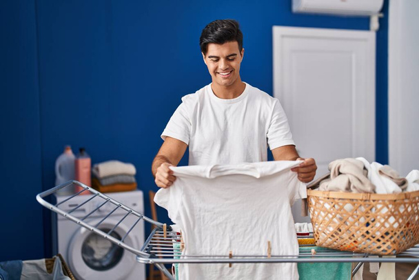 Młody Latynos uśmiecha się pewnie wieszając ubrania na sznurku w pralni - Zdjęcie, obraz