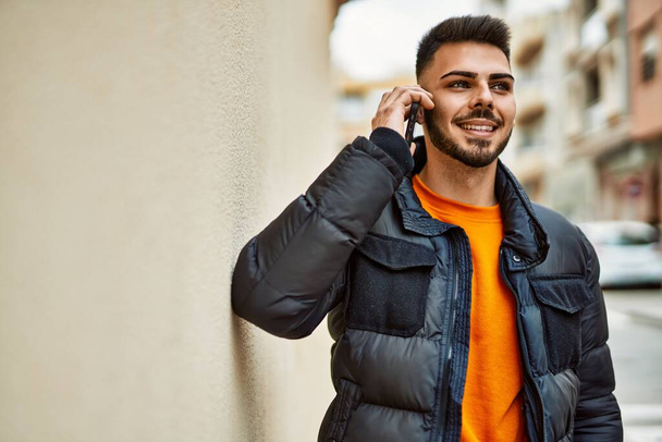 Jóképű, szakállas spanyol férfi, boldogan és magabiztosan mosolyog a városban, télikabátban beszél a telefonban. - Fotó, kép