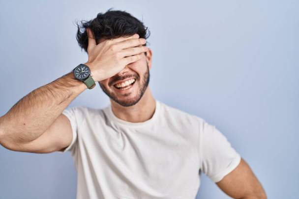 Hispanic man with beard standing over white background smiling and laughing with hand on face covering eyes for surprise. blind concept.  - Foto, Imagen