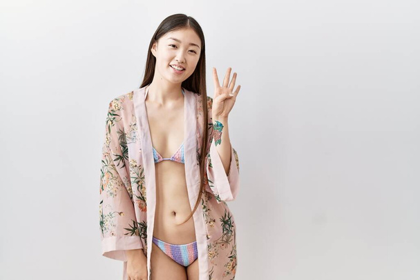 Молодая азиатка в бикини, в цветочном кимоно, показывает пальцами номер четыре, улыбаясь уверенно и счастливо..  - Фото, изображение