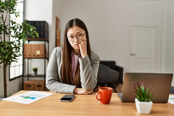 Młody chiński pracownik biznesu w stylu biznesowym siedzi na biurku w biurze myśląc patrząc zmęczony i znudzony problemami depresji z skrzyżowanymi ramionami.  - Zdjęcie, obraz