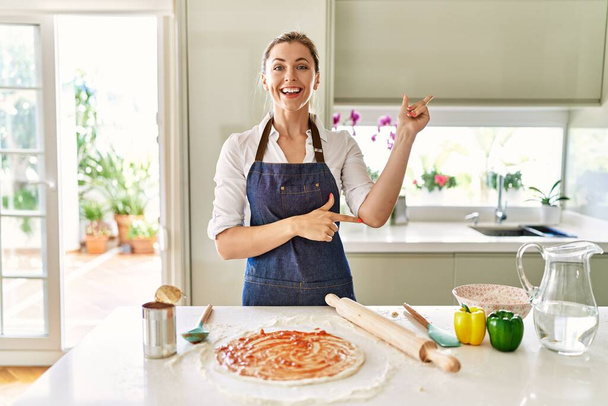 Красивая блондинка в фартуке готовит пиццу улыбаясь счастливо указывая рукой и пальцем в сторону  - Фото, изображение