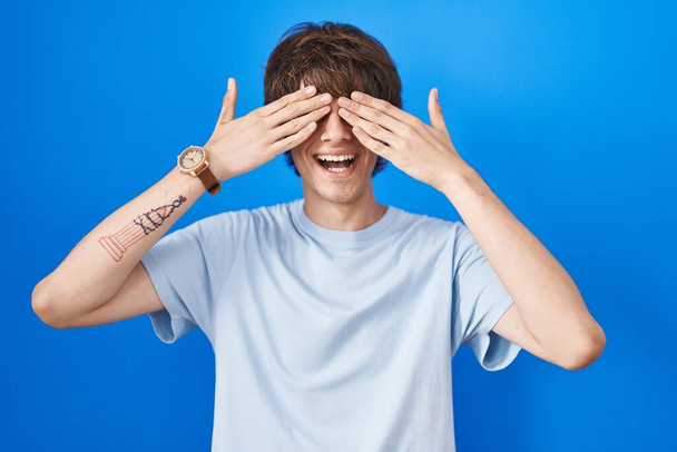 Hispanic young man standing over blue background covering eyes with hands smiling cheerful and funny. blind concept.  - Photo, Image