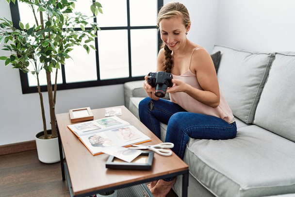 Young caucasian woman making photo book holding camera sitting on sofa at home - Photo, image