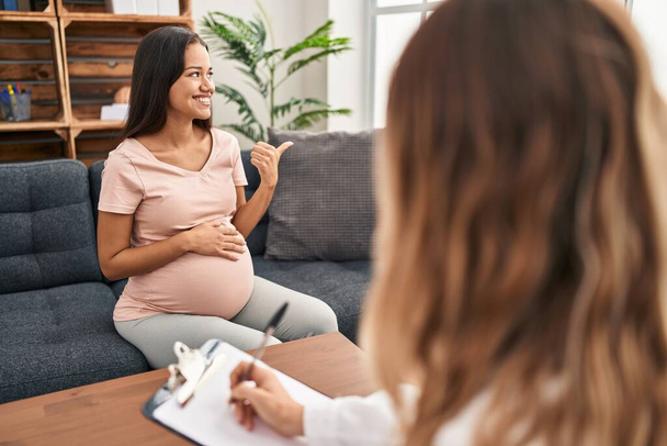 Terapi seansında gülümseyen ve gülümseyen genç hamile bir kadın başparmağıyla yan tarafa bakıyor..  - Fotoğraf, Görsel