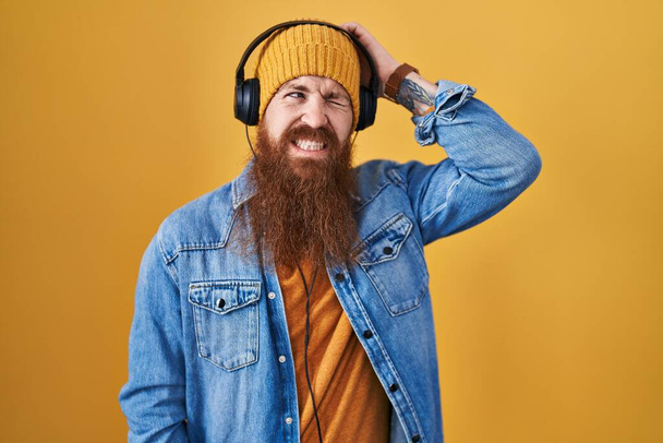 Caucasian man with long beard listening to music using headphones confuse and wondering about question. uncertain with doubt, thinking with hand on head. pensive concept.  - Foto, Imagen