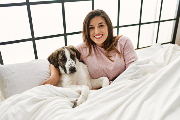 Mujer joven sonriendo confiado abrazando perro sentado en la cama en el dormitorio - Foto, imagen