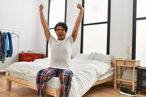 Young hispanic man waking up stretching arms sitting on the bed at bedroom. - Foto, Imagen