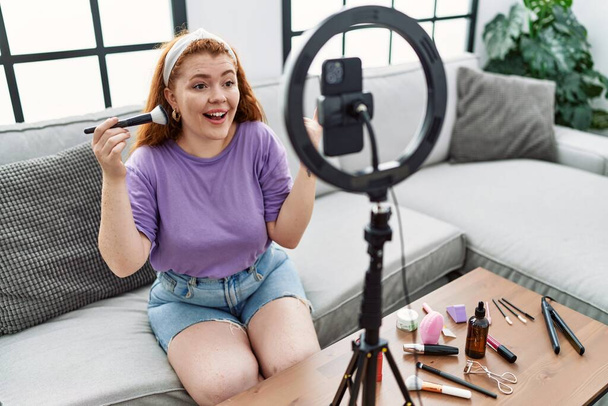 Mladá zrzka žena nahrávání make-up tutorial s smartphonem doma s úsměvem šťastný ukazující rukou a prstem na stranu  - Fotografie, Obrázek