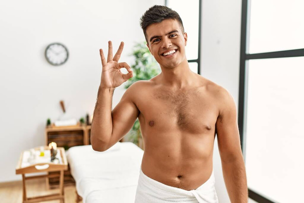 Jeune homme hispanique debout torse nu au centre de spa souriant positif faire ok signe avec la main et les doigts. expression réussie.  - Photo, image