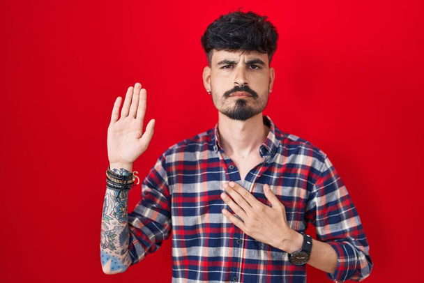 Young hispanic man with beard standing over red background swearing with hand on chest and open palm, making a loyalty promise oath  - Zdjęcie, obraz