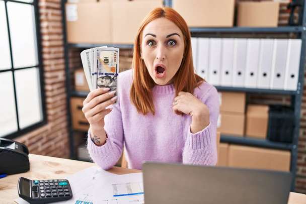 Młoda kobieta pracująca w małym biznesie e-commerce trzymająca pieniądze przerażona i zdumiona otwartymi ustami dla zaskoczenia, niedowierzanie twarz  - Zdjęcie, obraz