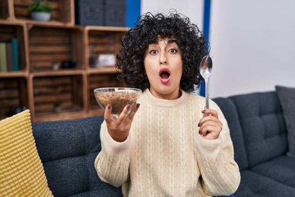 Young brunette woman with curly hair eating healthy whole grain cereals with spoon afraid and shocked with surprise and amazed expression, fear and excited face.  - Fotoğraf, Görsel
