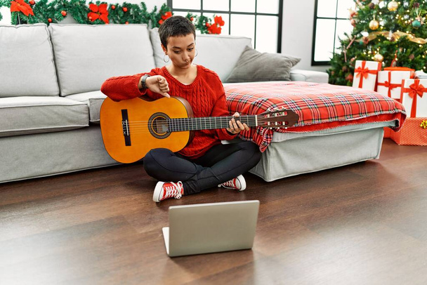 Hispanic woman with short hair learning classical guitar from online tutorial with angry face, negative sign showing dislike with thumbs down, rejection concept  - Foto, Imagem