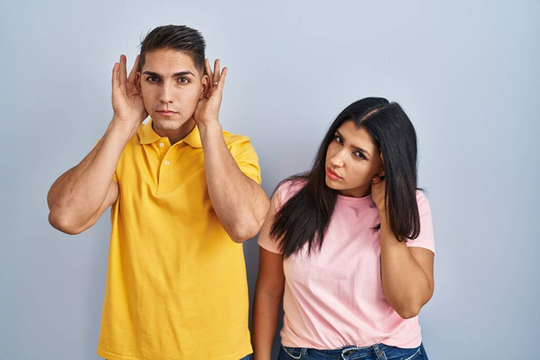 Νεαρό ζευγάρι στέκεται πάνω από απομονωμένο υπόβαθρο προσπαθεί να ακούσει και τα δύο χέρια σε χειρονομία αυτί, περίεργος για κουτσομπολιά. πρόβλημα ακοής, κωφός  - Φωτογραφία, εικόνα
