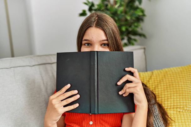 Adorable girl covering mouth with book at home - Fotoğraf, Görsel