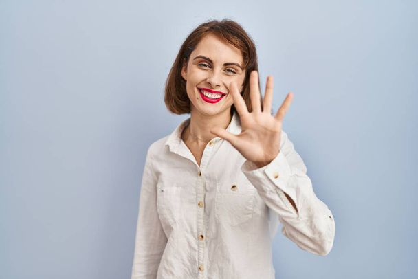 Young beautiful woman standing casual over blue background showing and pointing up with fingers number five while smiling confident and happy.  - Zdjęcie, obraz