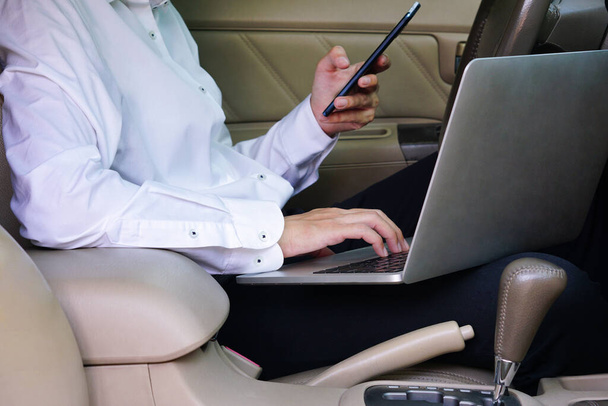 Επιχειρηματίας που εργάζεται σε φορητό υπολογιστή και κινητό τηλέφωνο, ενώ κάθεται στη θέση του οδηγού στο αυτοκίνητο. Έννοια τρόπου ζωής. - Φωτογραφία, εικόνα