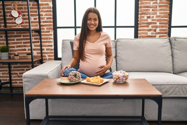 Mujer embarazada joven comiendo dulces en casa con aspecto positivo y feliz de pie y sonriendo con una sonrisa confiada mostrando los dientes  - Foto, Imagen