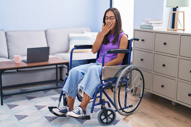 Νεαρή Ισπανίδα που κάθεται σε αναπηρική καρέκλα στο σπίτι και βαριέται χασμουριέται καλύπτοντας το στόμα με το χέρι. ανησυχία και υπνηλία.  - Φωτογραφία, εικόνα