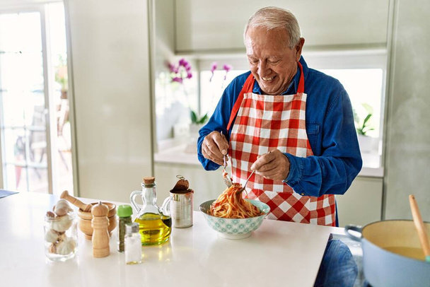 Senior man lacht vol vertrouwen tomatensaus met spaghetti mixen in de keuken - Foto, afbeelding
