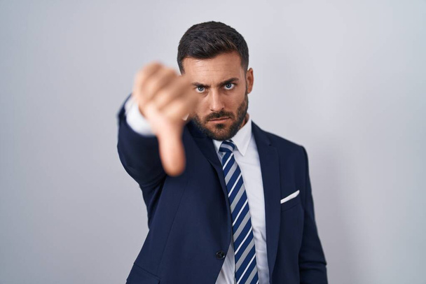 Jóképű spanyol férfi öltönyt és nyakkendőt visel, boldogtalannak és dühösnek tűnik, elutasítást és negatívat mutat hüvelykujjal lefelé. rossz kifejezés.  - Fotó, kép