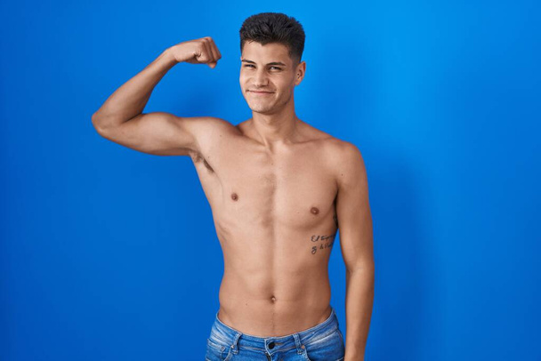 Young hispanic man standing shirtless over blue background strong person showing arm muscle, confident and proud of power  - Foto, Imagem