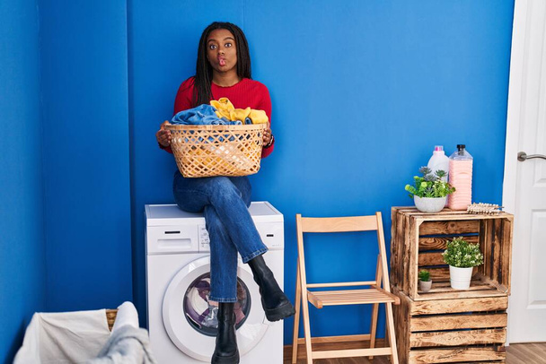 Young african american with braids holding laundry basket sitting on washing machine making fish face with mouth and squinting eyes, crazy and comical.  - Fotoğraf, Görsel