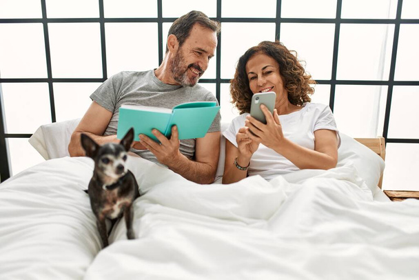 Середньовіччя його іспанська пара читає книгу і використовує смартфон. Лежачи на ліжку з собакою вдома
. - Фото, зображення