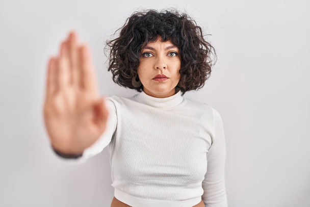 Hispanic woman with curly hair standing over isolated background doing stop sing with palm of the hand. warning expression with negative and serious gesture on the face.  - Foto, afbeelding