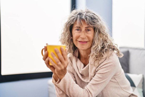 Middle age woman drinking coffee sitting on sofa at home - Фото, зображення