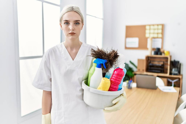 Young caucasian woman wearing cleaner uniform holding cleaning products cleaning office thinking attitude and sober expression looking self confident  - Foto, immagini