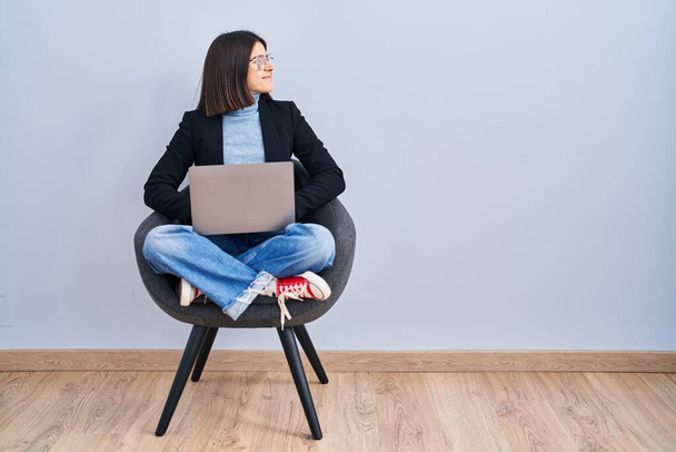 Νεαρή ισπανόφωνη γυναίκα κάθεται στην καρέκλα χρησιμοποιώντας φορητό υπολογιστή κοιτάζοντας προς τα πλάγια, να χαλαρώσετε προφίλ ποζάρουν με φυσικό πρόσωπο και αυτοπεποίθηση χαμόγελο.  - Φωτογραφία, εικόνα