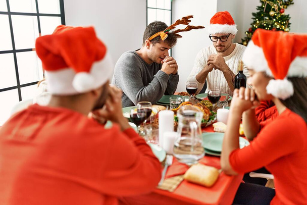 Groupe de jeunes célébrant Noël priant pour la nourriture assis sur la table à la maison. - Photo, image