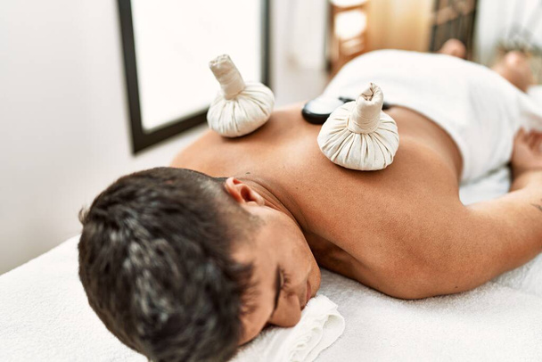 Joven hombre hispano relajado teniendo masaje de espalda usando piedras negras y bolsas tailandesas en el centro de belleza - Foto, Imagen